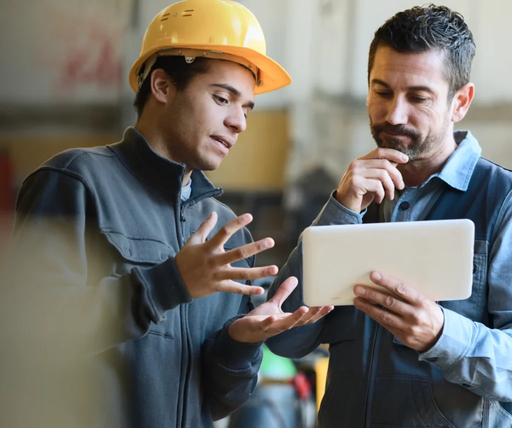 A man in a yellow hard hat explains something to another man holding a tablet.